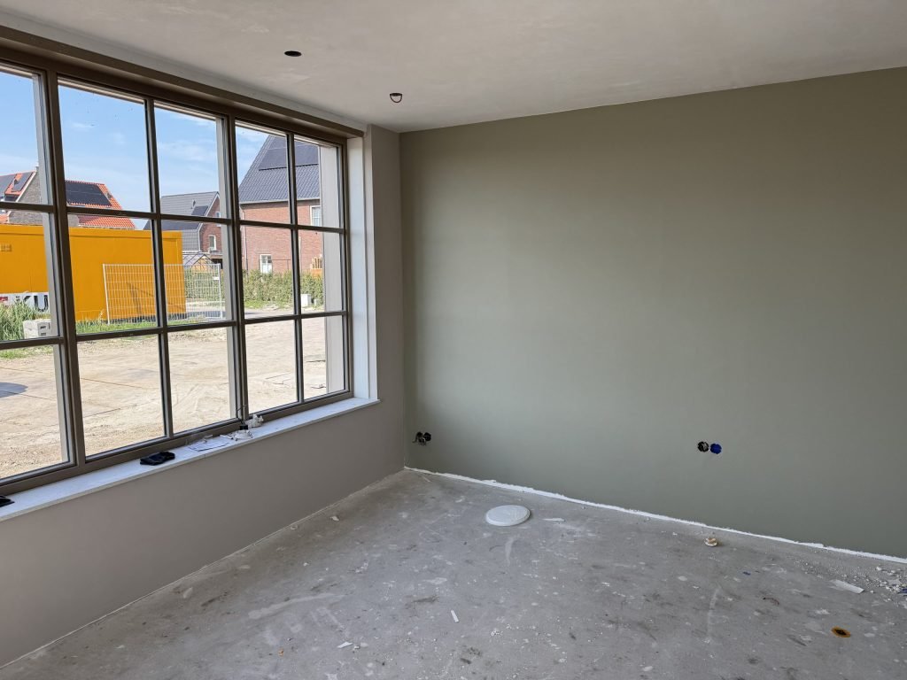 Gesausde wand in een grijsgroene kleur naast een groot raam met zicht op nieuwbouw