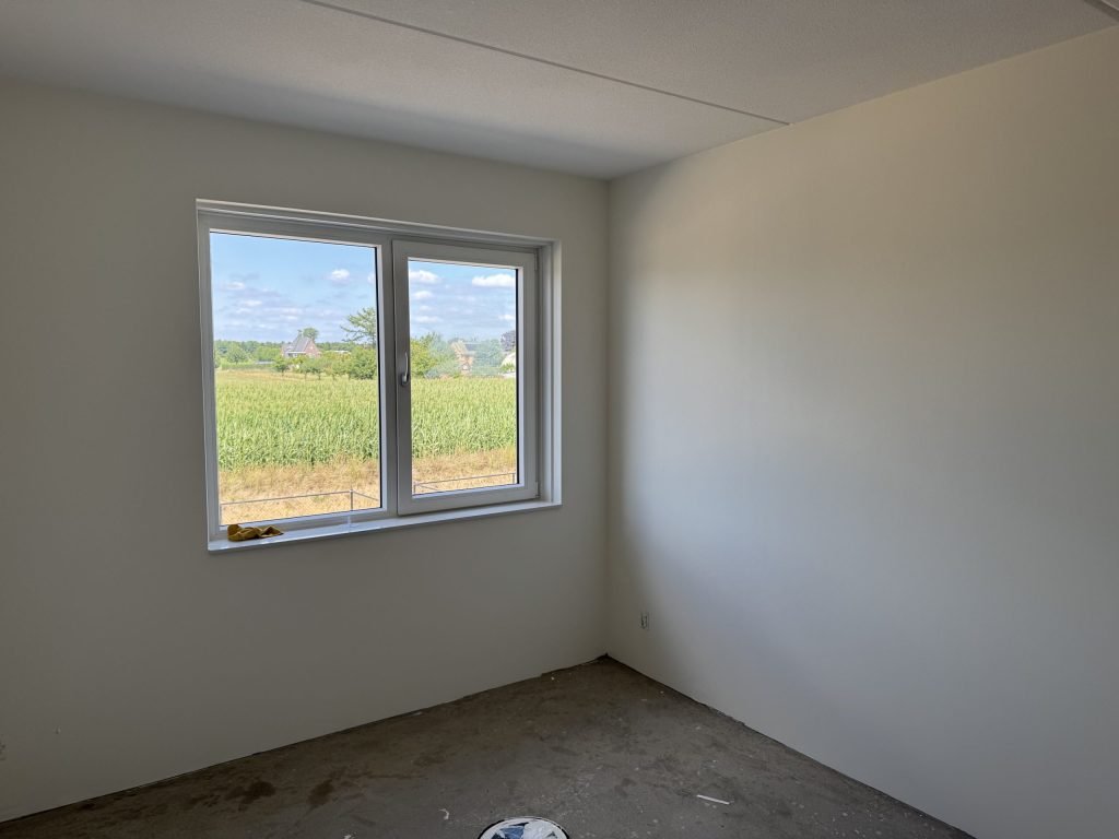 Kale slaapkamer in een nieuwbouwhuis met uitzicht op een maisveld en boerderijen